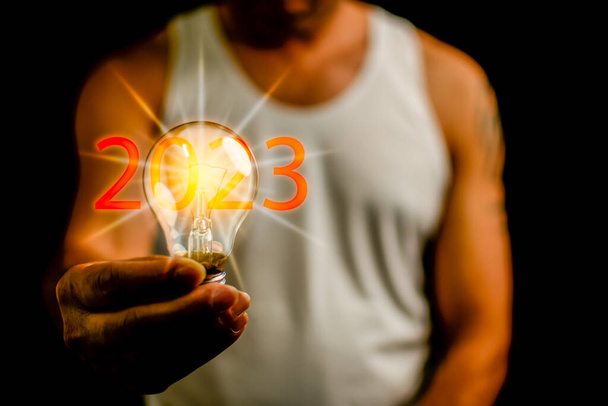 2023 Новогодняя концепция: Человек с лампочкой - это концепция стартапа, который управляет глобальным бизнесом. Яркие огни дают надежду малому бизнесу. Крупный план, скопировать пространство, черный размытый фон - Фото, изображение