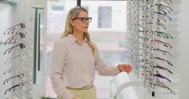 Portrait of a female optician or optometrist smiling and standing with her arms crossed between shelves in her shop. Confident mature woman and owner choosing or selling glasses at optics store. - Filmagem, Vídeo