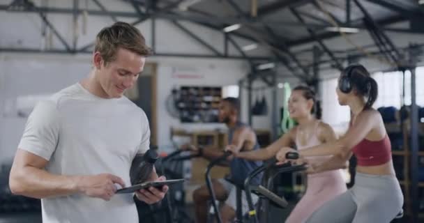 Coach using a digital tablet in a gym while training a group fitness class. Portrait of a muscular young male trainer smiling while browsing health and wellness data online during an aerobics workout. - Filmagem, Vídeo