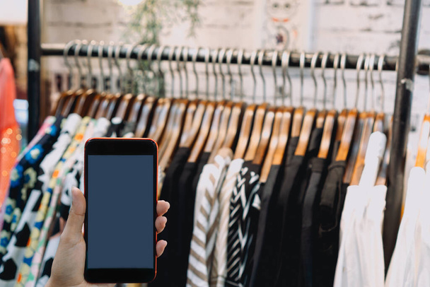 Γυναίκα στο κατάστημα ειδών ένδυσης και κρατώντας τηλέφωνο, Ρούχα σε ξύλινο φόντο κρεμάστρες, ψώνια σε απευθείας σύνδεση έννοια. - Φωτογραφία, εικόνα