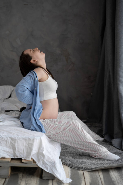 Beautiful pregnant female having a back pain while sitting on a bed in her apartment. Pregnancy and backache. pregnant lady touching pain zone on back sitting in the bedroom. - Foto, Bild