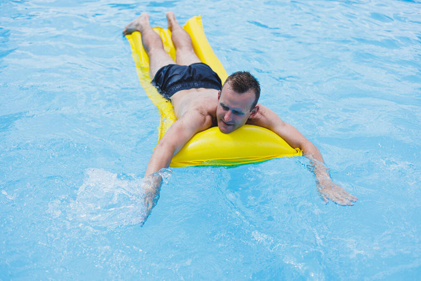 A young man in shorts enjoys the water park floating in an inflatable big ring on a sparkling blue pool smiling at the camera. Summer vacation. - Fotó, kép