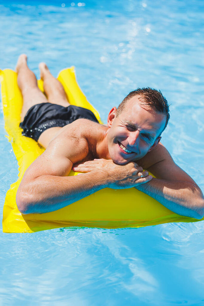 Egy rövidnadrágos fiatalember élvezi, hogy a vízi park egy felfújható nagy gyűrűben lebeg egy csillogó kék medencében, mosolyogva a kamerába. Nyári szünet. - Fotó, kép