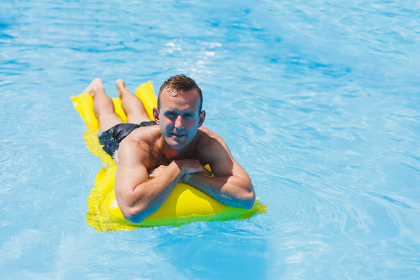 A young man in shorts enjoys the water park floating in an inflatable big ring on a sparkling blue pool smiling at the camera. Summer vacation. - 写真・画像