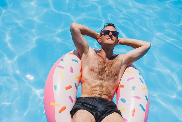 Ένας νεαρός άνδρας με γυαλιά ηλίου και σορτς χαλαρώνει σε ένα φουσκωτό ντόνατ στην πισίνα. Καλοκαιρινές διακοπές - Φωτογραφία, εικόνα