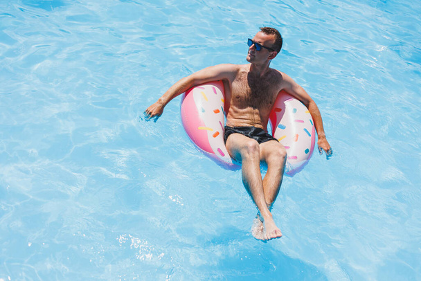 Ein junger Mann in Sonnenbrille und kurzer Hose entspannt auf einem aufblasbaren Donut im Pool. Sommerferien - Foto, Bild