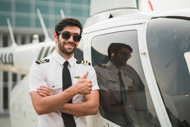 Portrait d'un pilote d'hélicoptère optimiste debout avec un avion et souriant en regardant la caméra. Bel homme beau et confiant au hangar extérieur. métier de porte-avions. Prêt à voler. - Photo, image