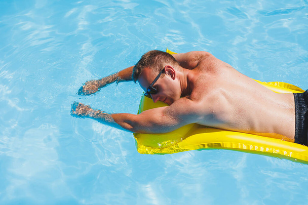 Un joven atractivo en gafas de sol y pantalones cortos se relaja en un colchón amarillo inflable en la piscina. Vacaciones de verano - Foto, Imagen