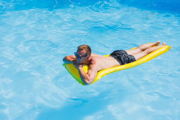 Attractive young man in sunglasses and shorts is relaxing on an inflatable yellow mattress in the pool. Summer vacation - Fotoğraf, Görsel