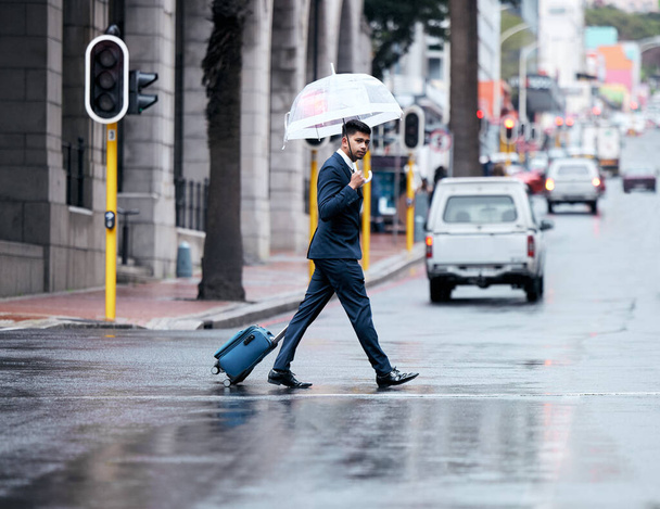 a young businessman walking across a street with a suitcase while holding an umbrella on a rainy day in the city. - Φωτογραφία, εικόνα