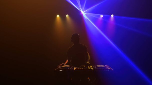 DJ - Silhouette of the disc jockey mixing music. Lights and laser of discotheque. Blue and gold colored lights - Zdjęcie, obraz