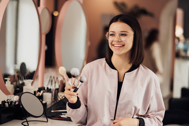 Πορτρέτο του νεαρού εύθυμη γυναίκα Make Up Artist Στέκεται στο σαλόνι ομορφιάς, Καυκάσια γυναίκα σε γυαλιά κρατώντας βούρτσες στο χώρο εργασίας - Φωτογραφία, εικόνα