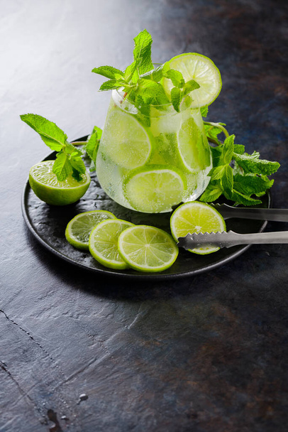 Mojito cocktail, mint and lime slices on a dark background. Summer cocktail mojito and ice tongs on a black plate. Top view. Copy space - Φωτογραφία, εικόνα