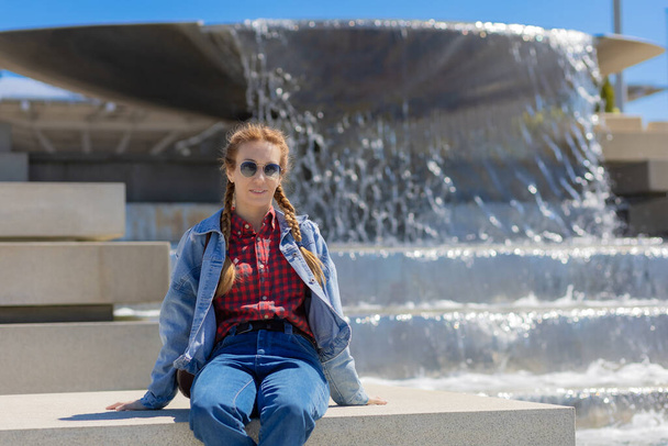 Une femme en chemise à carreaux et tenue en denim est assise sur un trottoir près d'une fontaine de la ville en cascade un jour de printemps ensoleillé. - Photo, image