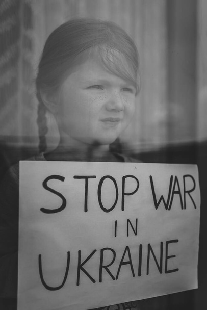 Child at the window and calls No War. Poor girl protesting war conflict raises banner with inscription Stop war in Ukraine. Stop russian aggression. Evacuation of civilians. Black and white photo. - Photo, Image