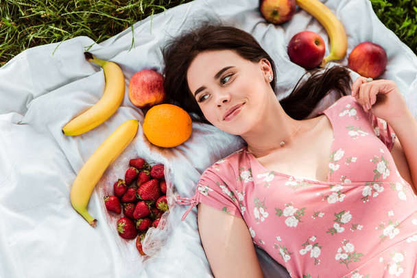 Woman lying on white bedsheet, looking at the camera, smiling. Lady have a rest in the city park, green grass lawn. A lot of fruits around. Travel, vacation, picnic set - Foto, afbeelding