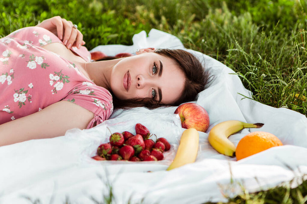 Woman lying on white bedsheet, looking at the camera, smiling. Lady have a rest in the city park, green grass lawn. A lot of fruits around. Travel, vacation, picnic set - Photo, image