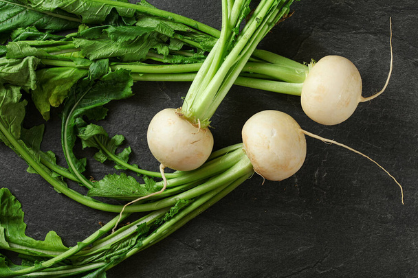 Three white radish bulbs with green leaves on black slate like board, view from above - 写真・画像
