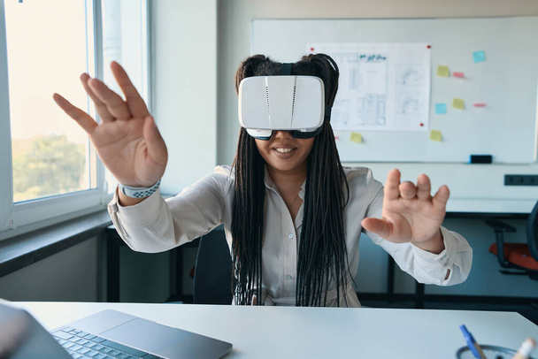 Smiling joyous female office worker sitting at office desk in VR headset - 写真・画像