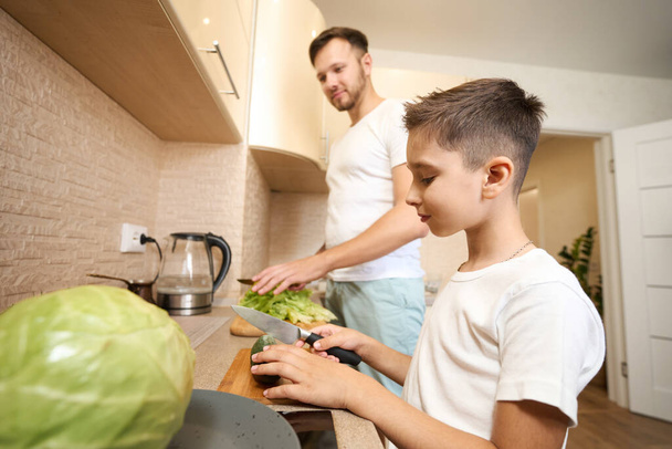 Focused Caucasian boy slicing cucumber with knife on cutting board next to his father - Valokuva, kuva