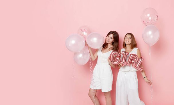 Portrait of happy two young women, dressed in a white, holdings a balloons, over pink background. - Foto, Imagen