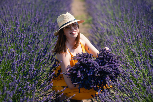 A young woman in a straw hat with a bunch of lavender flowers in a field among rows of lavender. - Valokuva, kuva