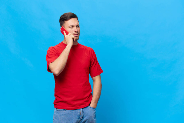 Раздраженный загорелый красавчик в красной футболке получает плохой финансовый отчет от телефонного звонка, позирующего изолированно на синем фоне. Копировать макет космического баннера. Концепция образа жизни онлайн-людей - Фото, изображение