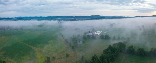 An aerial view from above the fog as it covers the landscape, fields, farms and small towns in the early morning light. - 写真・画像