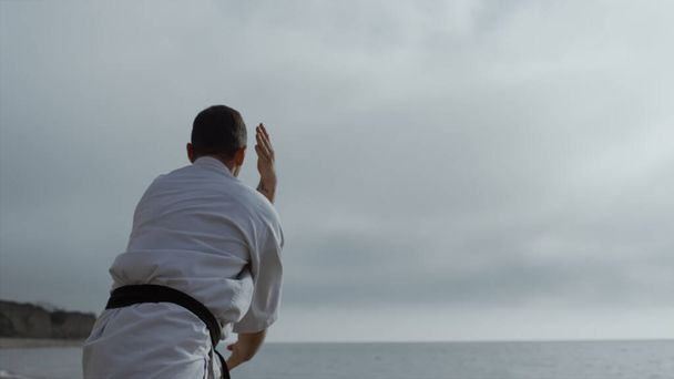Sporty man practicing karate fighting standing beach under gray sky. Focused sportsman training martial technique cloudy morning outdoors. Bearded athlete workout wearing kimono with black belt. - Zdjęcie, obraz
