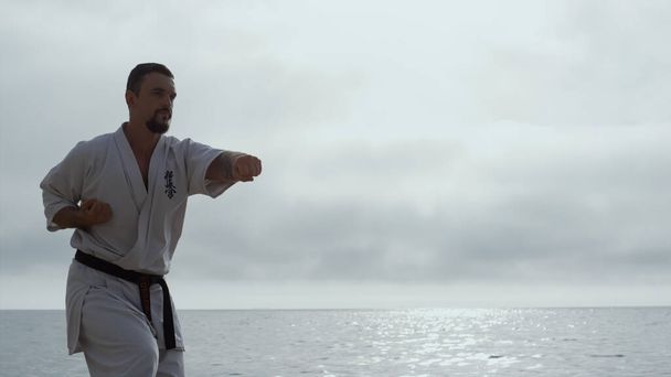 Serious sportsman learning martial arts cloudy day on seashore. Bearded man practicing fight skills against gray sky. Strong focused fighter training hands punches outdoors. Sport lifestyle concept. - Foto, Imagem