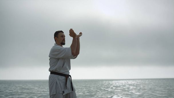 Taekwondo man training physical strength near calm sea. Bearded sportsman practicing martial arts wearing kimono overcast weather. Focused strong fighter making hands exercises workouting outdoors. - 写真・画像