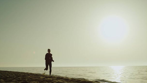 Silueta sportovkyň běžící na pobřeží před krásným západem slunce. Atletická dívka jogging písečné pláže v blízkosti klidné moře venku. Fit sportovní žena cvičení v přírodě letní večer. Zdravé cvičení. - Fotografie, Obrázek