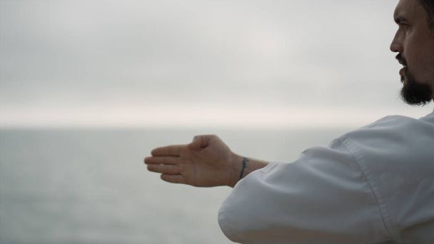Винищувач карате тренує техніку бойових мистецтв на морському пляжі хмарний день крупним планом. Бородатий чоловік займається літнім ранком у білому кімоно. Сильний спортсмен практикує удари руками. Спортивна концепція
. - Фото, зображення