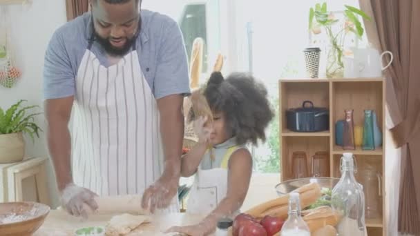 Famiglia afroamericana con padre che indossa grembiule rotolando farina trebbia per cucinare con figlia insieme in cucina a casa, genitore e bambino che preparano il cibo con divertimento e giocoso. - Filmati, video