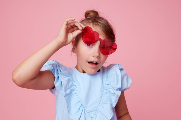 Ritratto di bambina carina sorpresa con gli occhiali da sole a forma di cuore. Bambino a bocca aperta che si diverte isolato su sfondo rosa. Guardando la telecamera. Wow faccia divertente - Foto, immagini