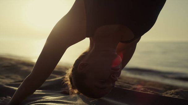 Yoga-Frau steht nach unten gerichteten Hund praktiziert svanasana bei Sonnenuntergang aus nächster Nähe. Schlankes, sportliches Mädchen, das an einem Sommertag an der Sandküste Flexibilität trainiert. Konzentrierte Dame Gesicht in der Nähe ruhigen Ozeanwellen Nahaufnahme. - Foto, Bild