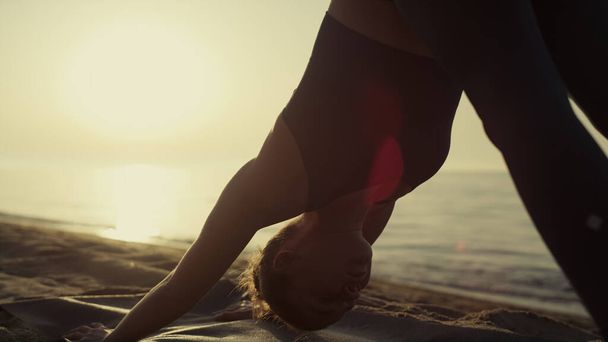 Fit sportswoman bending body standing yoga asana on sandy beach close up. Yogi woman practicing down face dog pose at sunset outside. Slim young girl training strength on seacoast summer evening. - Photo, Image