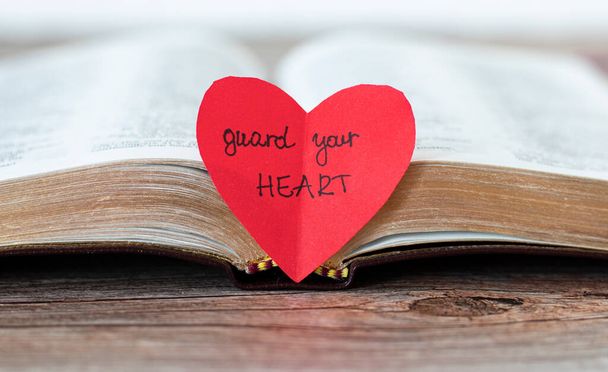 Punainen sydän käsin kirjoitetuilla sanoilla "Vartioi sydäntäsi", raamatullinen tekstijae ja avoin Raamattu-kirja kultaisilla sivuilla puupöydällä. Sananlaskut 4: 23. Lähemmäs. - Valokuva, kuva