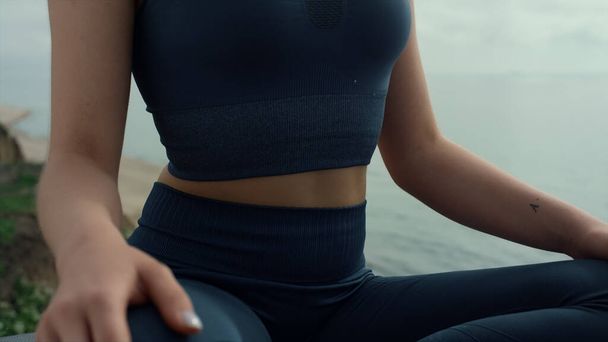 Fit pretty girl practicing pranayama on beach hill close up. Calm woman meditating in lotus pose closing eyes. Closeup sporty young body sitting on seacoast wearing black sportswear. Healthy lifestyle - Fotó, kép