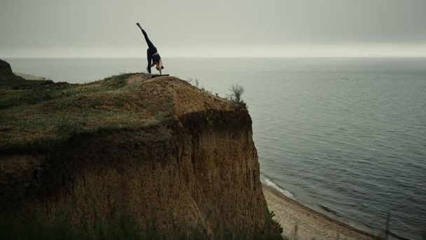 Młoda dziewczyna podnosi stopę do nieba stojąc na wysokim wzgórzu. Elastyczna kobieta rozciąga ciało pochylając się do ziemi pochmurny letni dzień. Sportowa pani ćwiczy jogę poza treningiem gimnastyka ćwiczenia na wzgórzu plaży.  - Zdjęcie, obraz