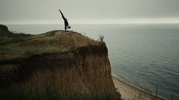 Bella donna flessibile piegando corpo a piedi praticando asana yoga sulla collina sulla spiaggia. Slim ragazza in piedi una gamba che si estende all'aperto. Donna sportiva che allena la pancetta indossando abbigliamento sportivo nero. Concetto sportivo. - Foto, immagini