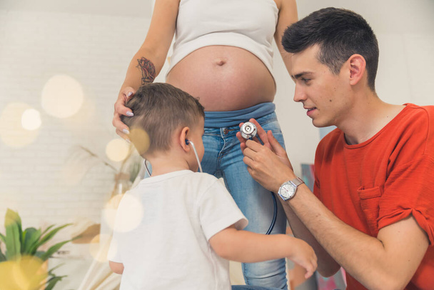 young man helping his son listen to the heartbeat of a baby in the belly of his wife, happy family. High quality photo - Foto, Imagen