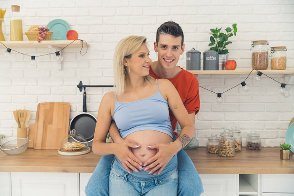 young Caucasian man hugging pregnant wife and touching her belly in the kitchen, medium shot happy marriage concept. High quality photo - Foto, Imagem