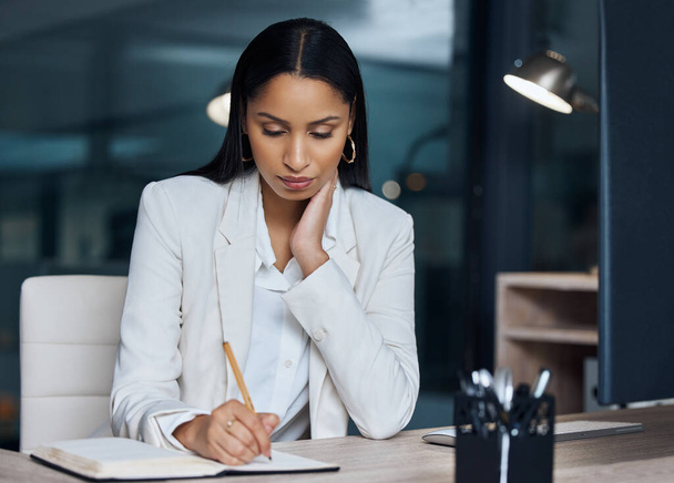 eine attraktive junge Geschäftsfrau, die nachts in ihrem Büro sitzt und beim Schreiben in ein Notizbuch nachdenklich wirkt. - Foto, Bild