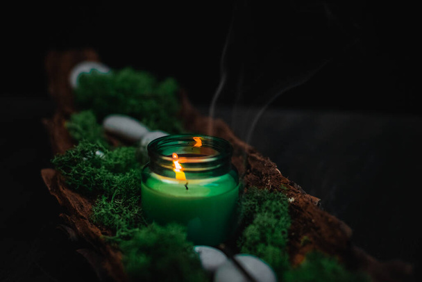 A white lit candle in a glass jar with flowing smoke, green moss and gray stones in the bark of a tree lies diagonally against a black background, close-up side view. Esoteric concept, dark style. - Photo, Image