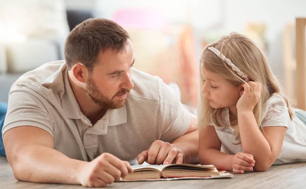 ένας νεαρός πατέρας και η κόρη διαβάζοντας ένα βιβλίο μαζί στο σπίτι. - Φωτογραφία, εικόνα