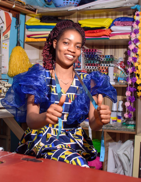 Eine glückliche afrikanische Schneiderin, Modedesignerin, Geschäftsfrau oder Unternehmerin, die sich hinsetzt, während sie in einer Schneiderei Daumen nach oben macht - Foto, Bild