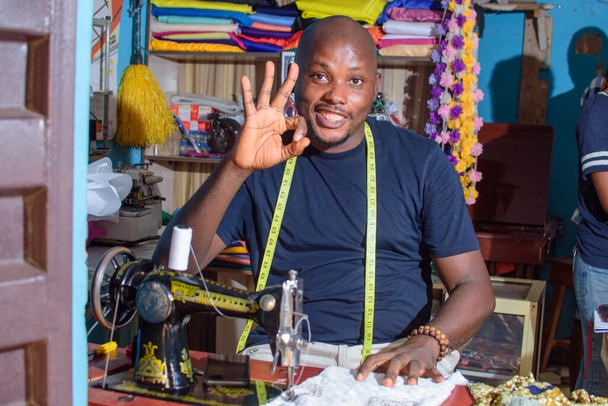 Un felice sarto maschio africano nigeriano, stilista o uomo d'affari che fa gesto mano eccellenza mentre fa vestito con una macchina da cucire in una sartoria - Foto, immagini