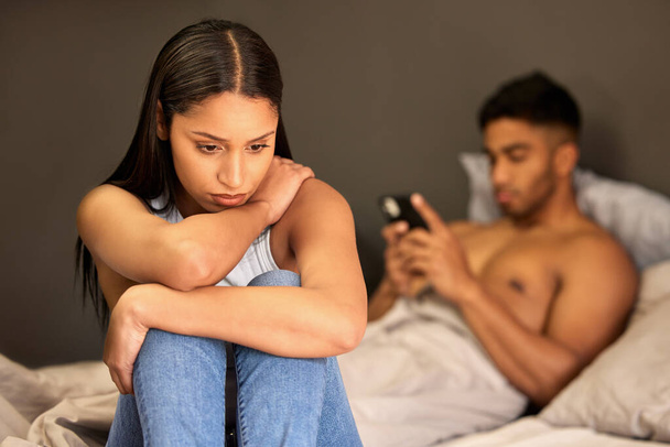 a young woman sitting on her bed and feeling upset while her boyfriend uses his cellphone behind her. - Zdjęcie, obraz