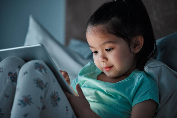 a little girl using a digital tablet while lying on her bed. - Foto, Bild